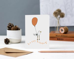 Seagul holding an orange baloon hand illustrated greeting card with handwritten happy birthday text in orange