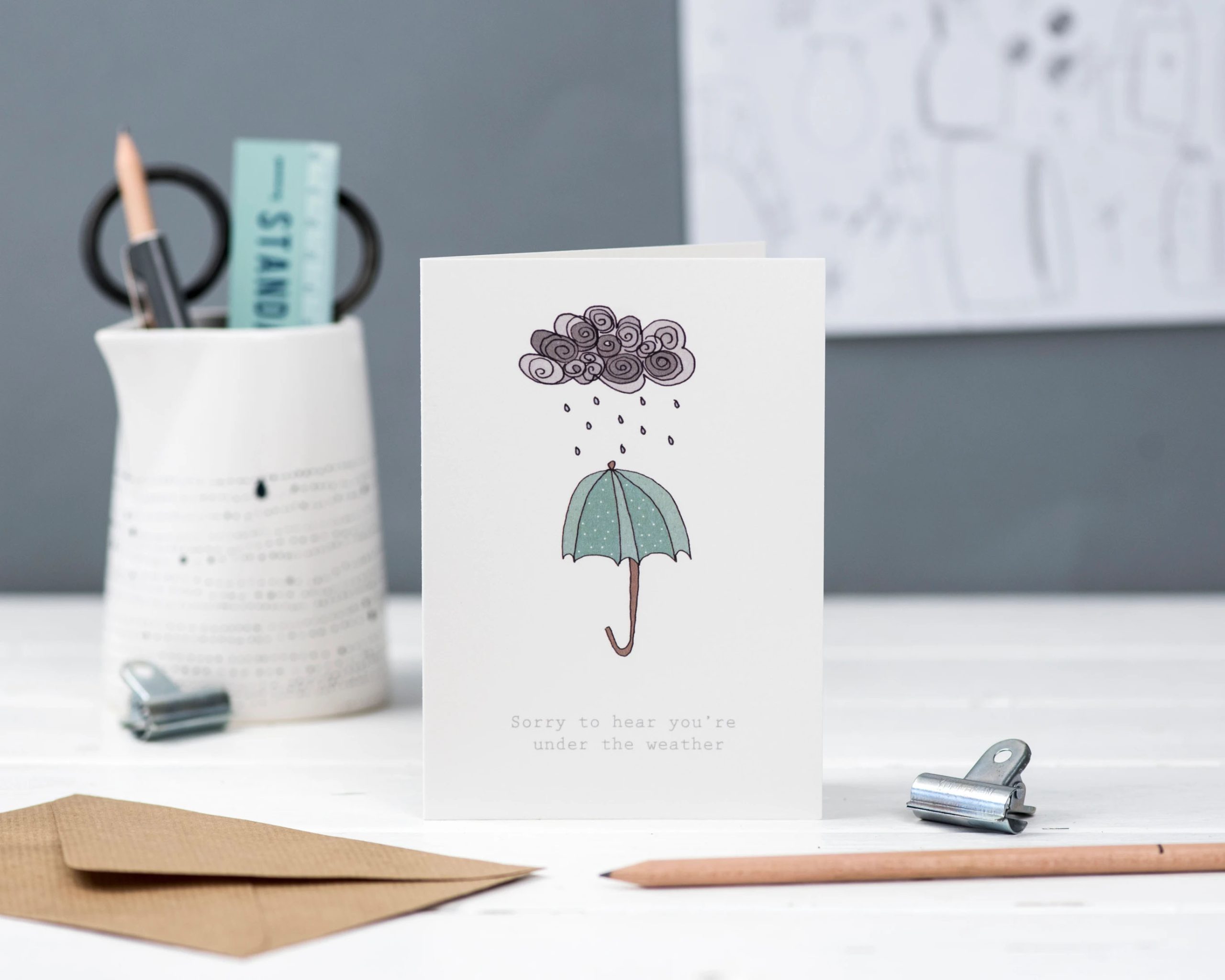 Get well soon greeting card with hand illustrated cloud raining on illustrated umbrella with text sorry to hear you're under the weather
